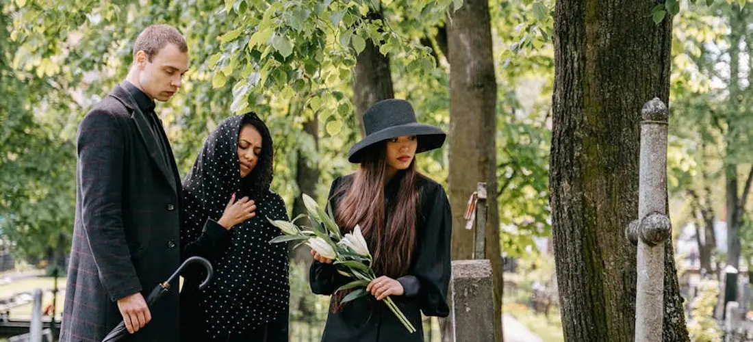 Three mourners in a cemetery dressed in understated dark clothing; a man in a coat, a woman in a black polka-dot outfit, and another woman wearing a wide-brim hat while holding flowers.