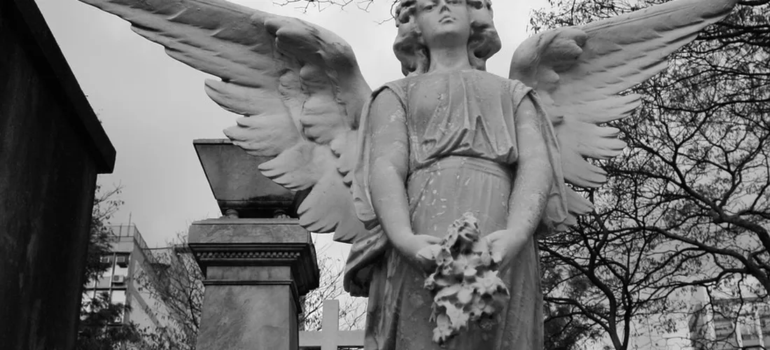 Grayscale photograph of an angel statue with outstretched wings holding a bouquet, standing among tombstones with bare trees in the background.