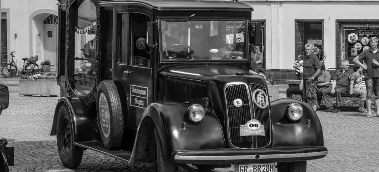 Black-and-white vintage hearse on a cobblestone street with onlookers and buildings in the background.