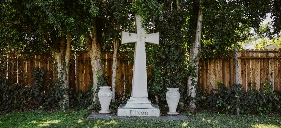 White cross tombstone flanked by two vases on a grassy plot with a wooden fence and trees in the background.
