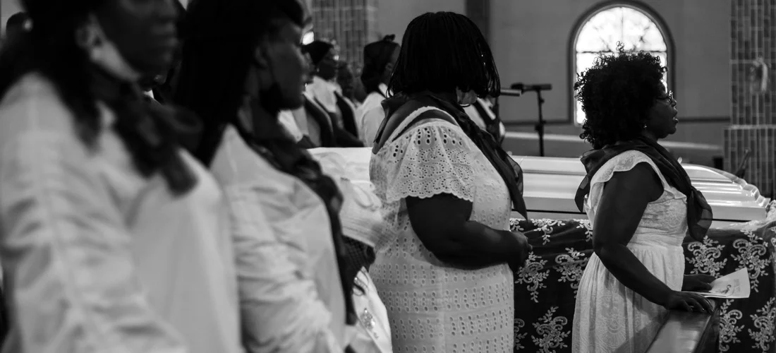 Black-and-white photo of a line of mourners in a church beside a closed casket, somber and respectful.