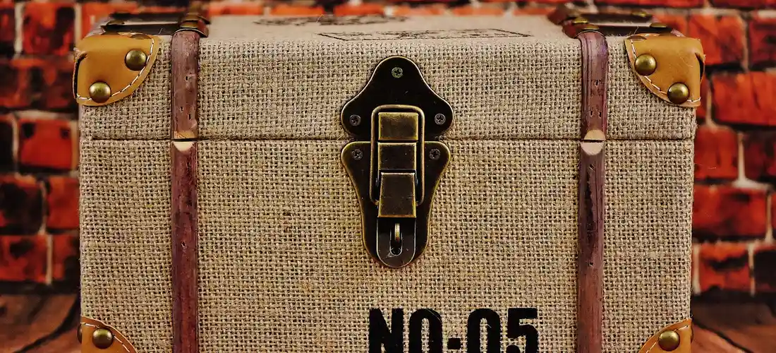 A burlap-covered casket-style box with brass corner protectors and a central latch, set against a red brick wall.