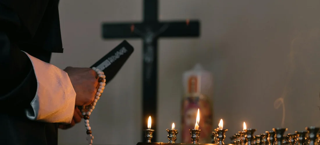 A clergyman holding a rosary in the foreground with lit candles and a cross in the background.
