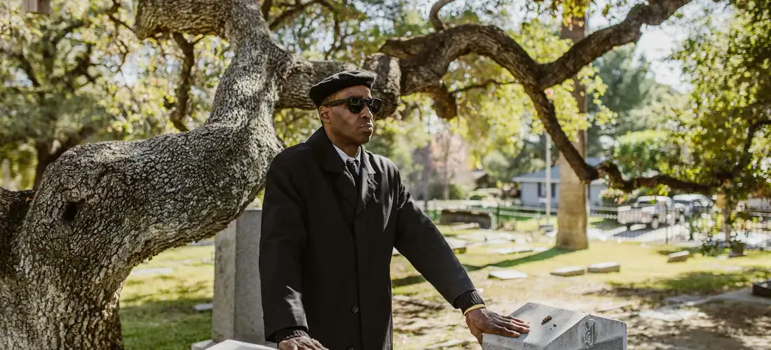 A man in a dark suit and hat stands beside a gravestone under a tree in a cemetery.