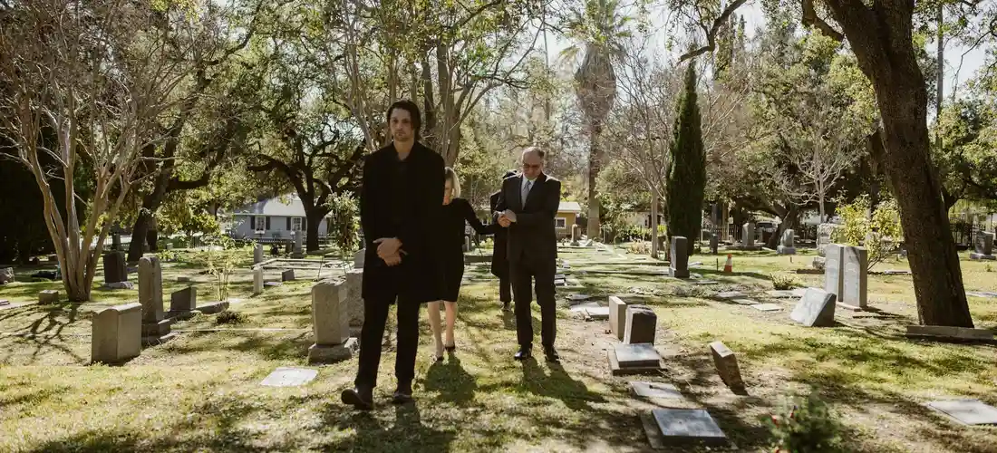 Two men in dark suits walk through a sunlit cemetery with gravestones and trees after a funeral.