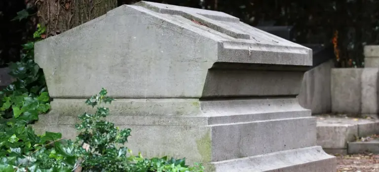 Weathered stone tomb in a cemetery surrounded by greenery.