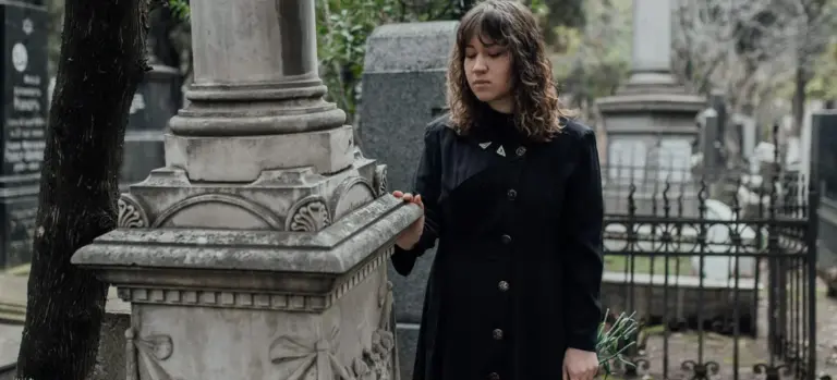 A woman in a dark coat stands beside a cemetery tombstone, representing financial planning for retirement and life events.