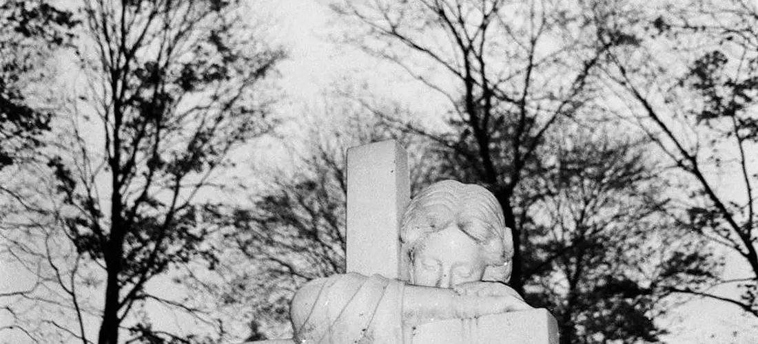 Black-and-white photo of a carved cemetery statue on a tombstone with bare trees in the background.
