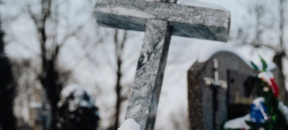 Close-up of a weathered cross-shaped tombstone in a cemetery with other headstones blurred in the background.