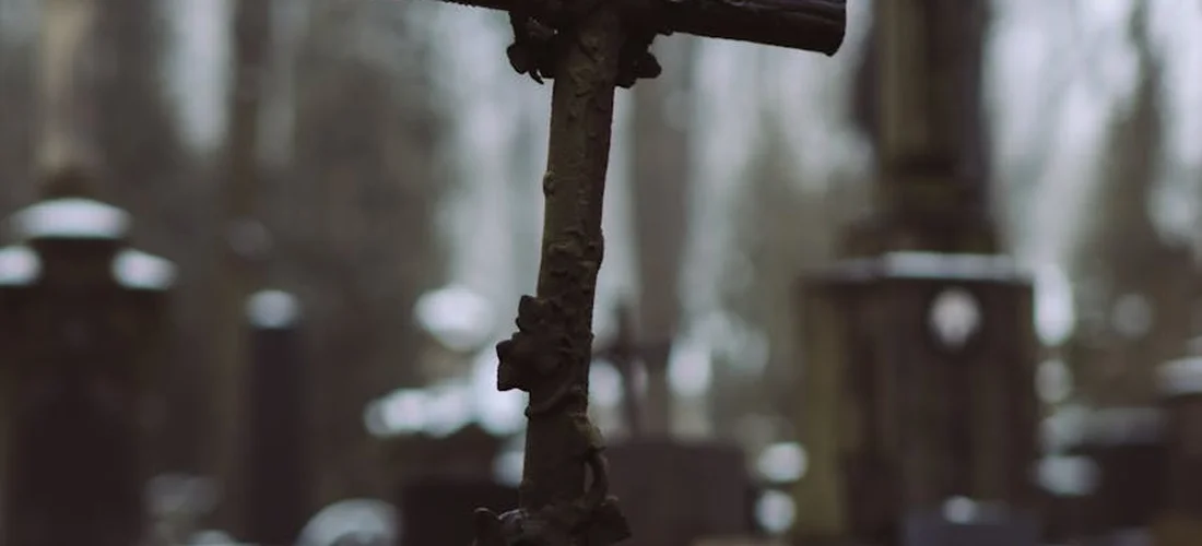 Close-up of a weathered iron cross in a graveyard with tombstones blurred in the background.