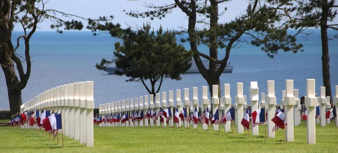 Rows of white grave markers adorned with small flags in a grassy cemetery, with trees and a blue sea in the background.