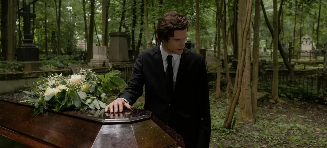 A man in a black suit standing by a closed casket in a cemetery, symbolizing funeral planning and paperwork.