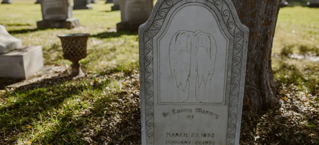 Gravestone with a carved relief of clasped hands in a cemetery, with grass and other headstones in the background.