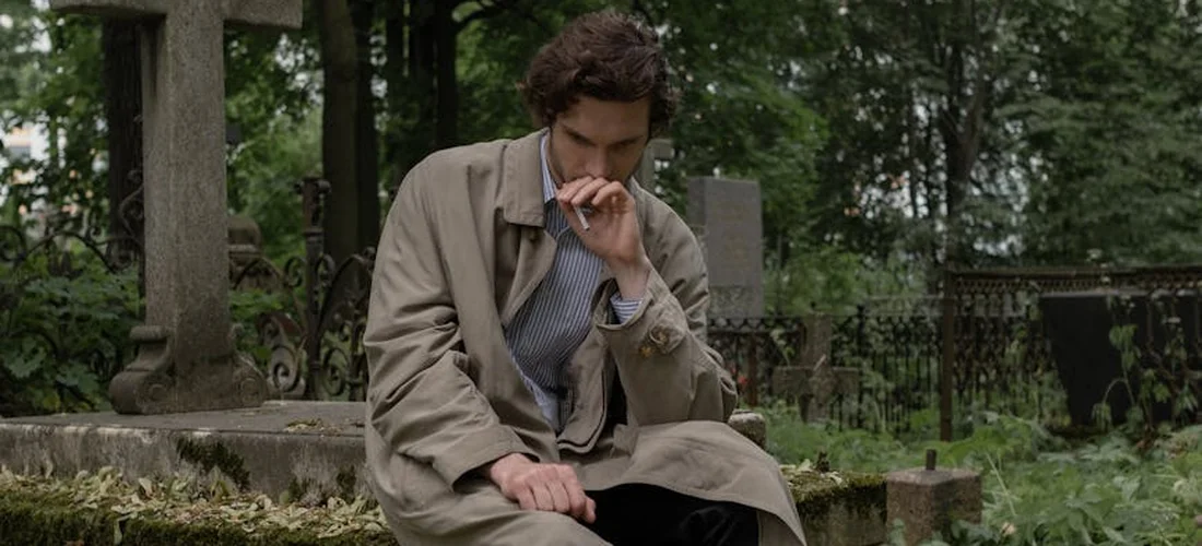 A man in a tan coat sits on a stone bench in a cemetery, head bowed with his hand near his mouth, surrounded by gravestones and green grass.