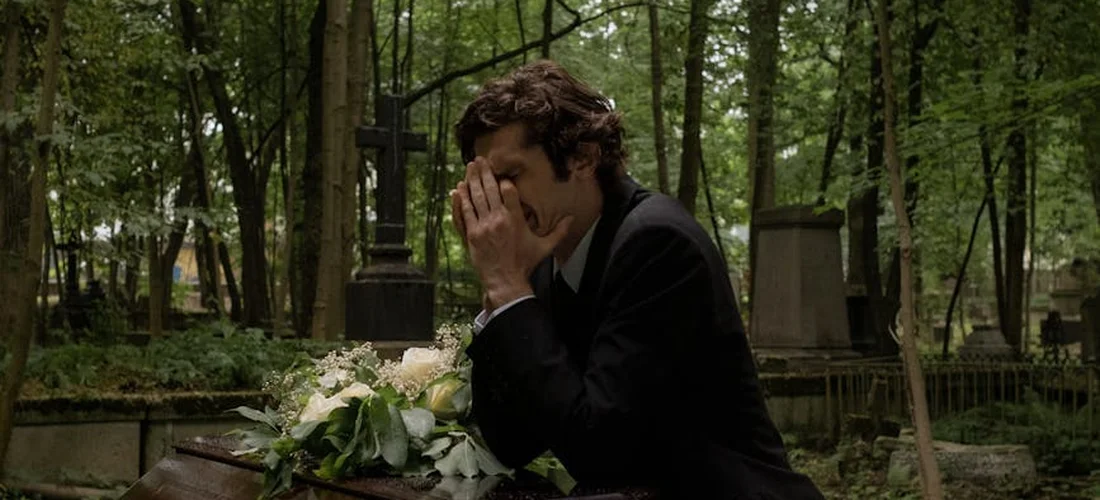 A person in a dark suit sits at a grave in a quiet cemetery, hands covering their face, with a bouquet of white flowers resting on the tomb.