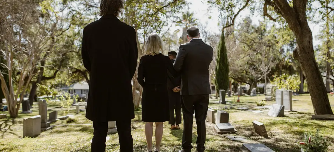 Group of mourners in dark attire standing in a sunlit cemetery, facing gravestones as the service begins.