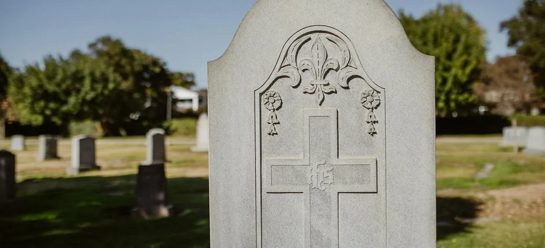 A carved tombstone with a cross in a cemetery, with other gravestones and trees in the blurred background