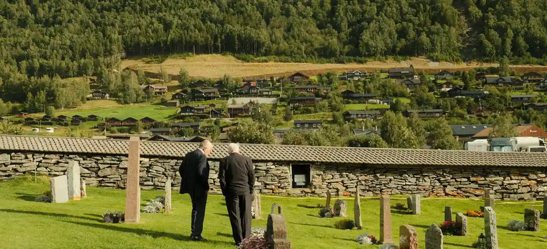 Two men in dark jackets walk through a cemetery with headstones, a stone wall, and a hillside village in the distance.