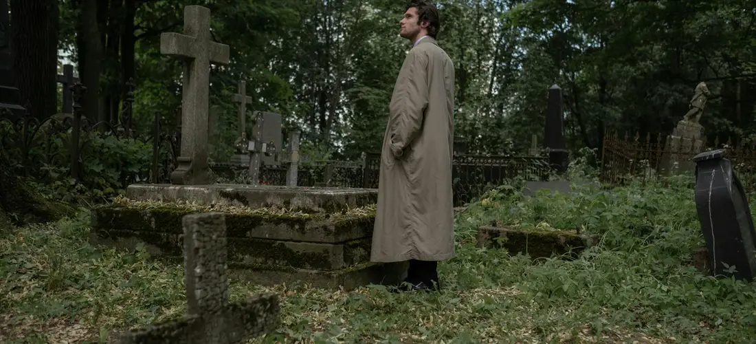 A person in a beige trench coat stands beside weathered gravestones in an overgrown cemetery, with trees in the background.
