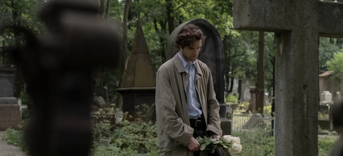 A man in a light jacket and tie stands in a cemetery, holding a bouquet of white flowers with tombstones in the background, looking down in a contemplative pose.