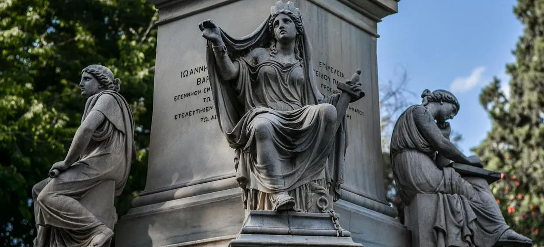 Three stone statues surrounding a tall cemetery monument outdoors, with greenery and a blue sky.