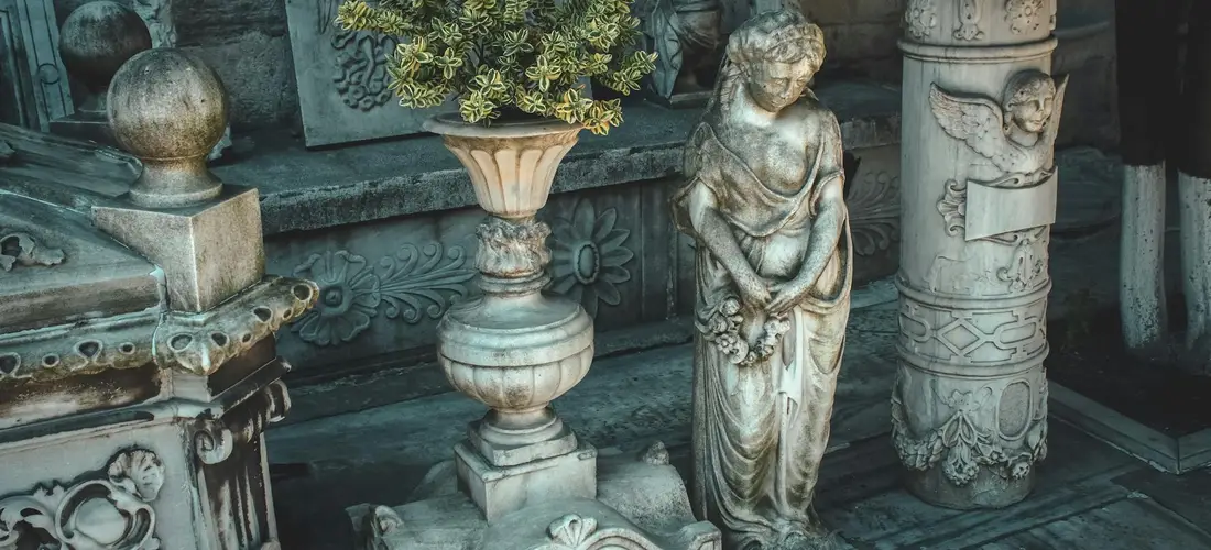 Stone cemetery scene featuring a pedestal urn and a standing statue among ornate memorials.