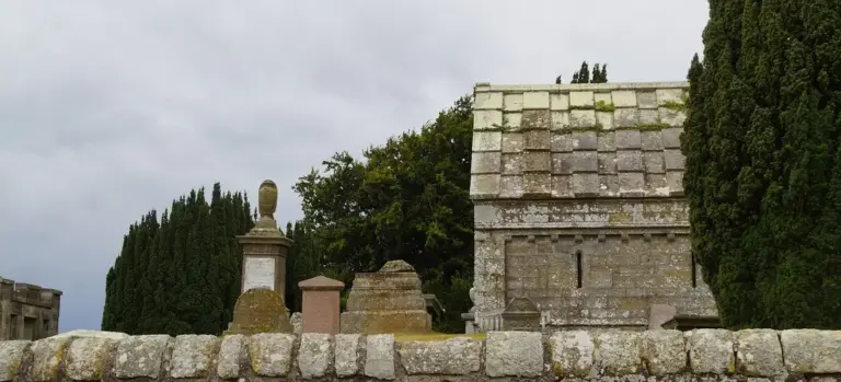 Gravestones and a stone mausoleum in a quiet cemetery with a cloudy sky, suggesting reverence and remembrance.