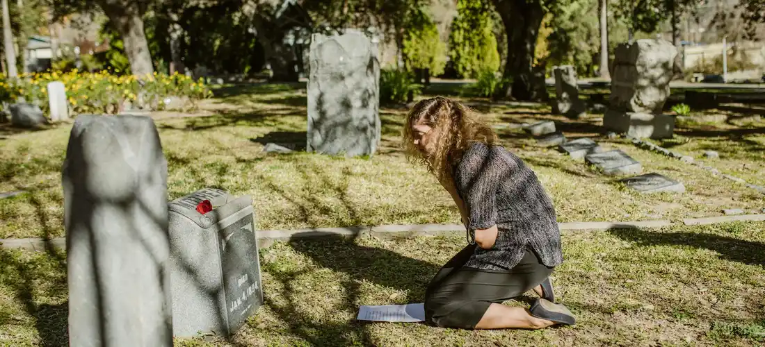 A person kneels beside a gravestone in a cemetery, with a small prayer book on the ground, as sunlight filters through the trees.