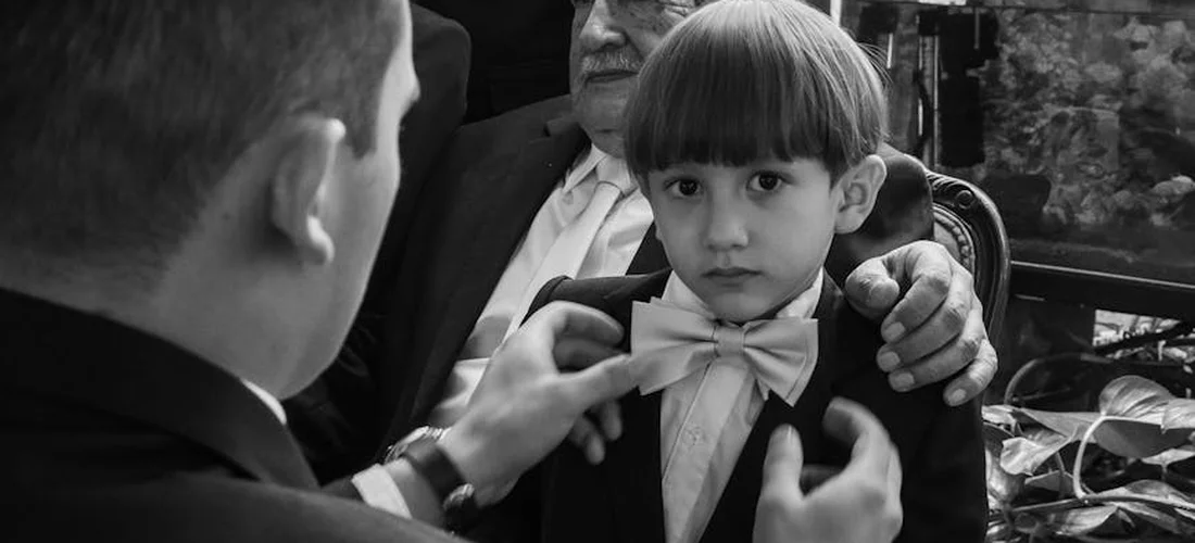 A young boy in a suit with a bow tie is being helped by an adult as his bow tie is adjusted