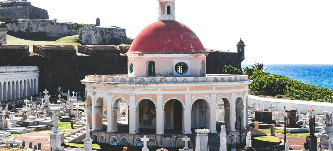 A domed seaside chapel at a coastal cemetery with numerous tombstones and the ocean in the background.