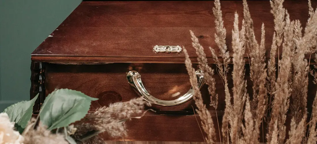 A wooden coffin with metallic handles, surrounded by dried ornamental grasses.