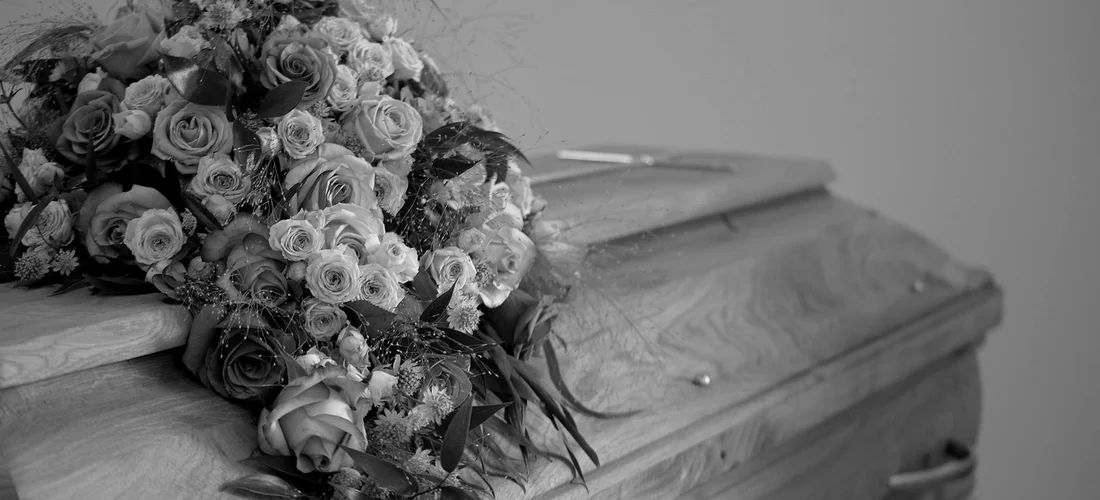 Black-and-white photo of a closed coffin topped with a large bouquet of roses and other flowers.