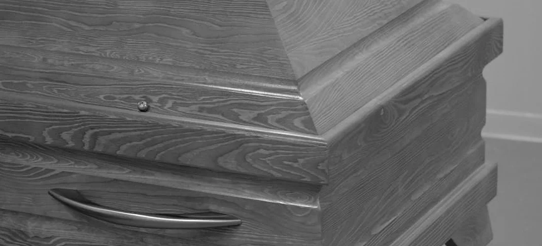 Close-up view of a wooden coffin with a metal handle, shown in grayscale