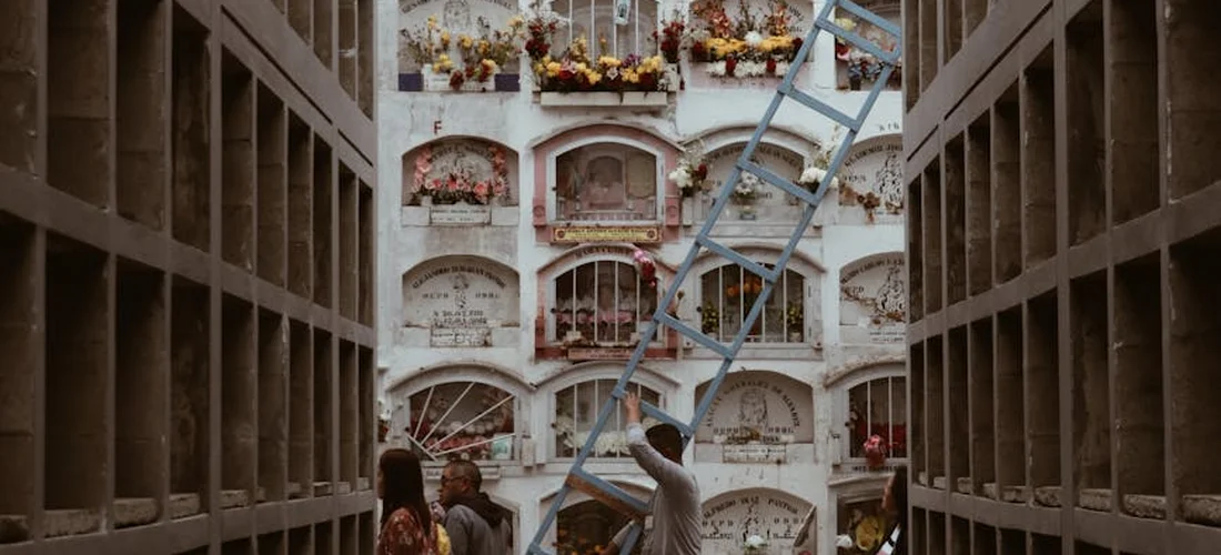 Interior view of a columbarium with rows of cremation niches, urns and floral arrangements; a person stands on a ladder among the niches.