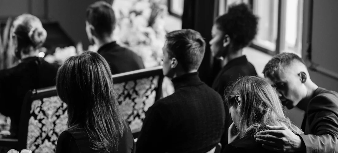 Mourners seated in pews during a funeral service in black and white