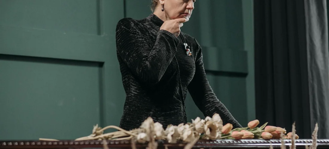 A grieving woman in a black dress stands beside a coffin adorned with flowers at a funeral.