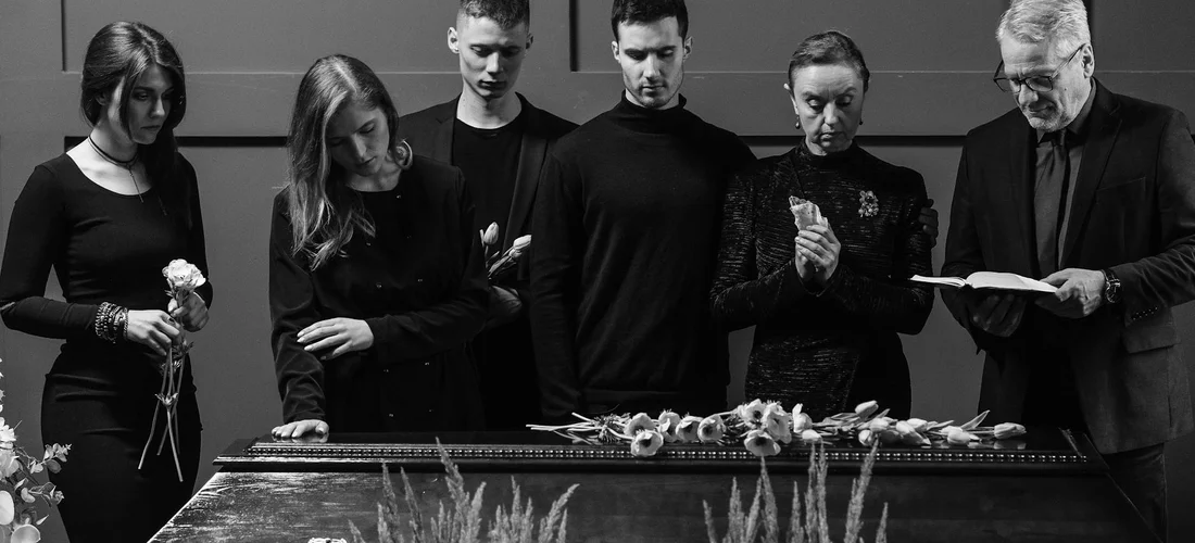Black-and-white photo of mourners in formal black attire standing beside a closed casket, conveying a traditional burial ceremony.