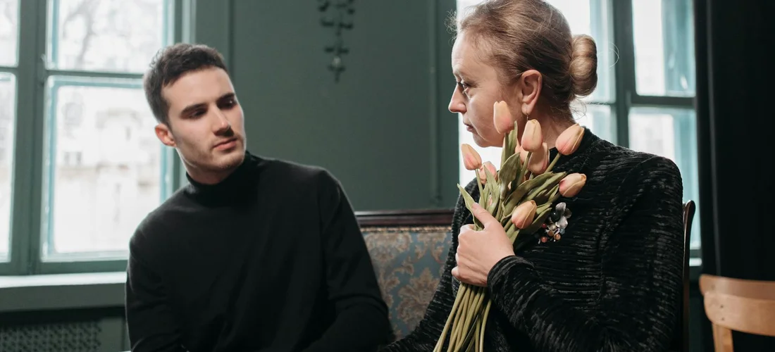 Two people in a softly lit room discuss memorial flowers; one person holds a bundle of dried blooms, illustrating a personal approach to DIY funeral arrangements.