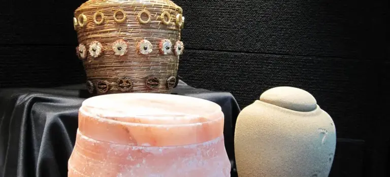 Two ceremonial urns or vessels on a dark fabric, one ornate brown urn and one smooth beige vase, illustrating cremation-related objects.