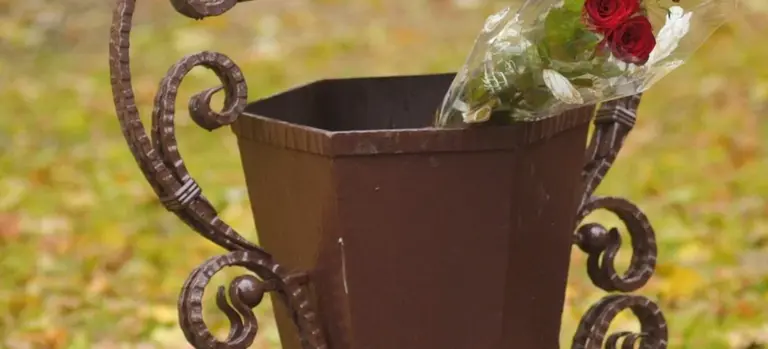 A brown metal cremation urn with a bouquet of red and white flowers placed inside, outdoors on a grassy, autumn-ground.