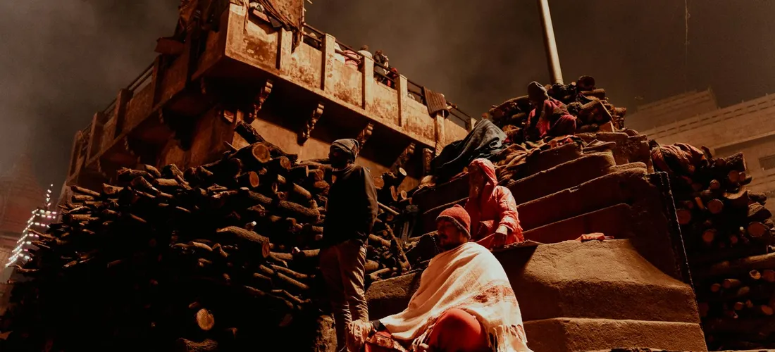 Mourners near a cremation platform with wooden logs stacked nearby during a cremation ceremony.