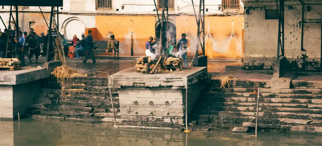 People at a traditional cremation ghat along a river, with stone platforms and ritual activity.