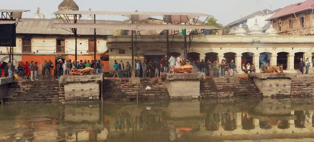 Riverside ghats with stone steps along the water and a crowd, with traditional buildings in the background.