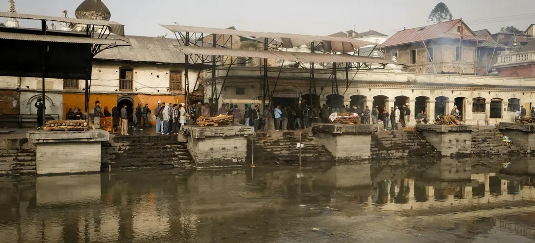 People gather along a riverfront cremation ghats with stone steps and wooden platforms, smoke rising as funeral rites are conducted.