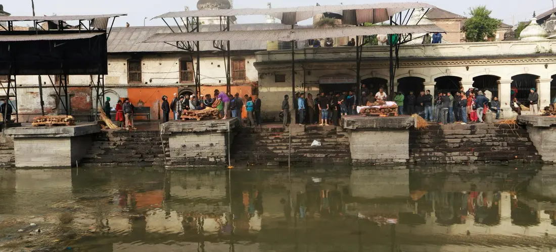 Riverside cremation ghats with stone platforms along a river and mourners gathered by the water, reflecting in the surface during a cremation ceremony.