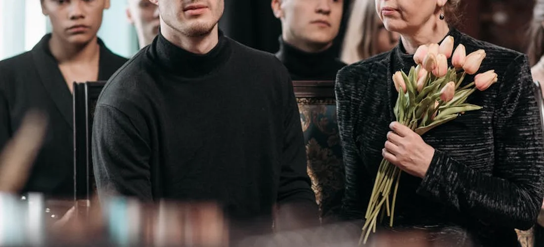 Group of mourners at a funeral service; a woman holding a bouquet of pink tulips stands beside a man in a black turtleneck, with other attendees in the background.