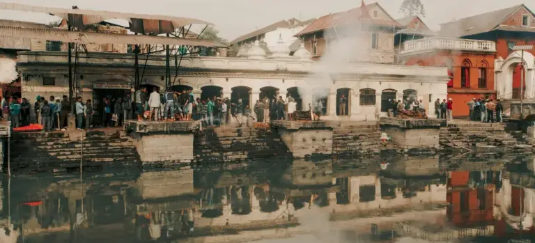 People gathered along a riverside cremation site as smoke rises from cremation pyres and buildings line the opposite bank.