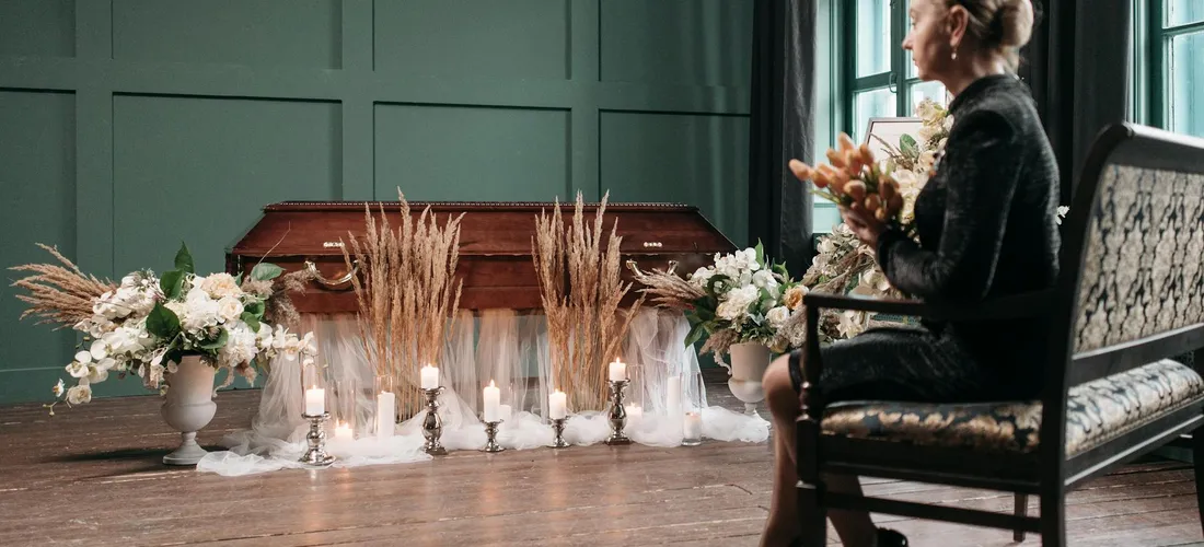 A funeral scene featuring a closed coffin, candles, and floral arrangements with a grieving person seated nearby.