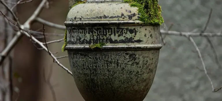 Close-up of a moss-covered, weathered metal cremation urn with faded inscriptions.