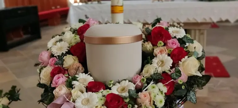Ceremonial white urn with a pink-gold band, encircled by a wreath of red, pink, and white roses and chrysanthemums.